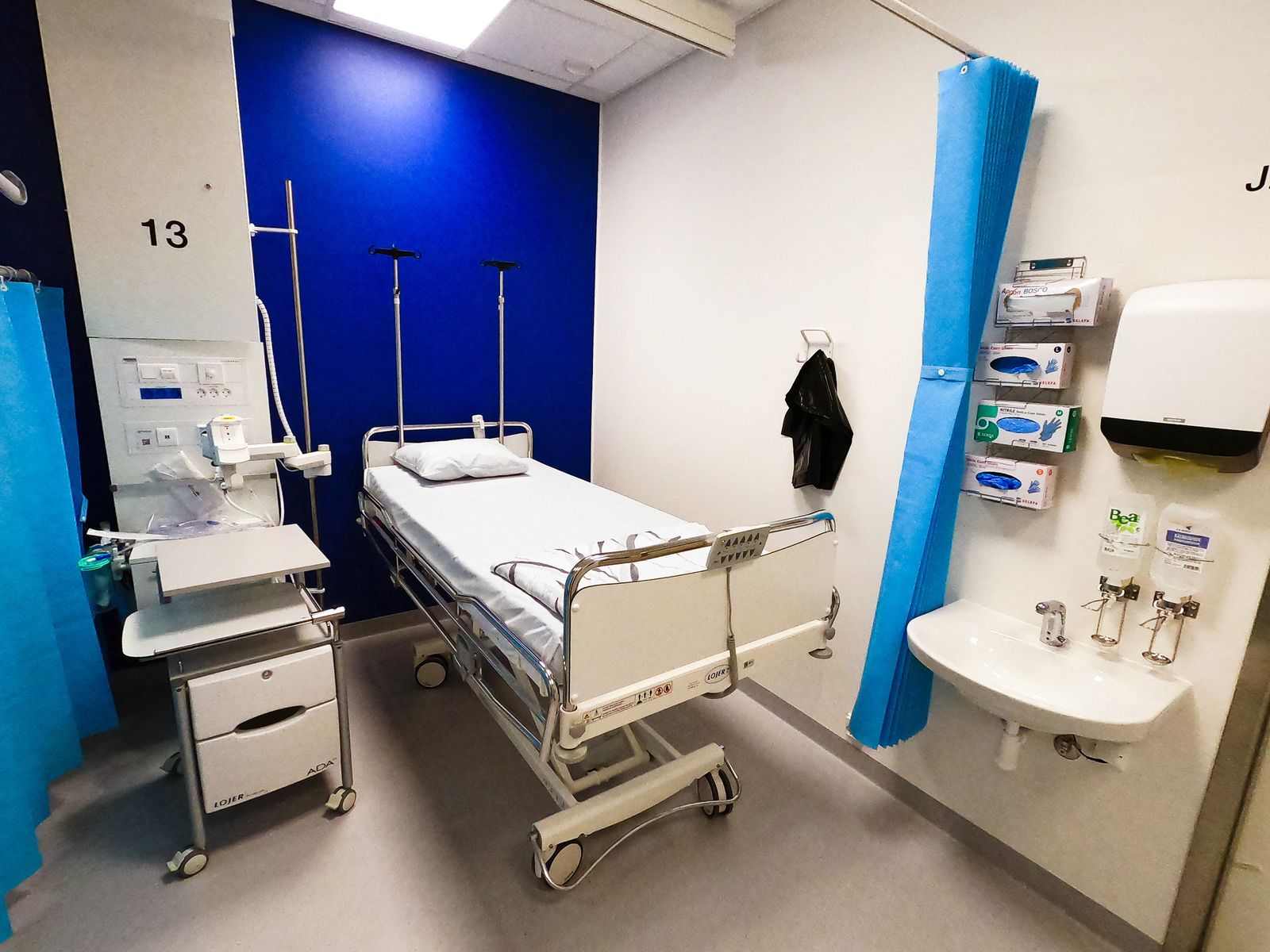A hospital bed in a single room beside a bedside cabinet and wall-mounted supplies